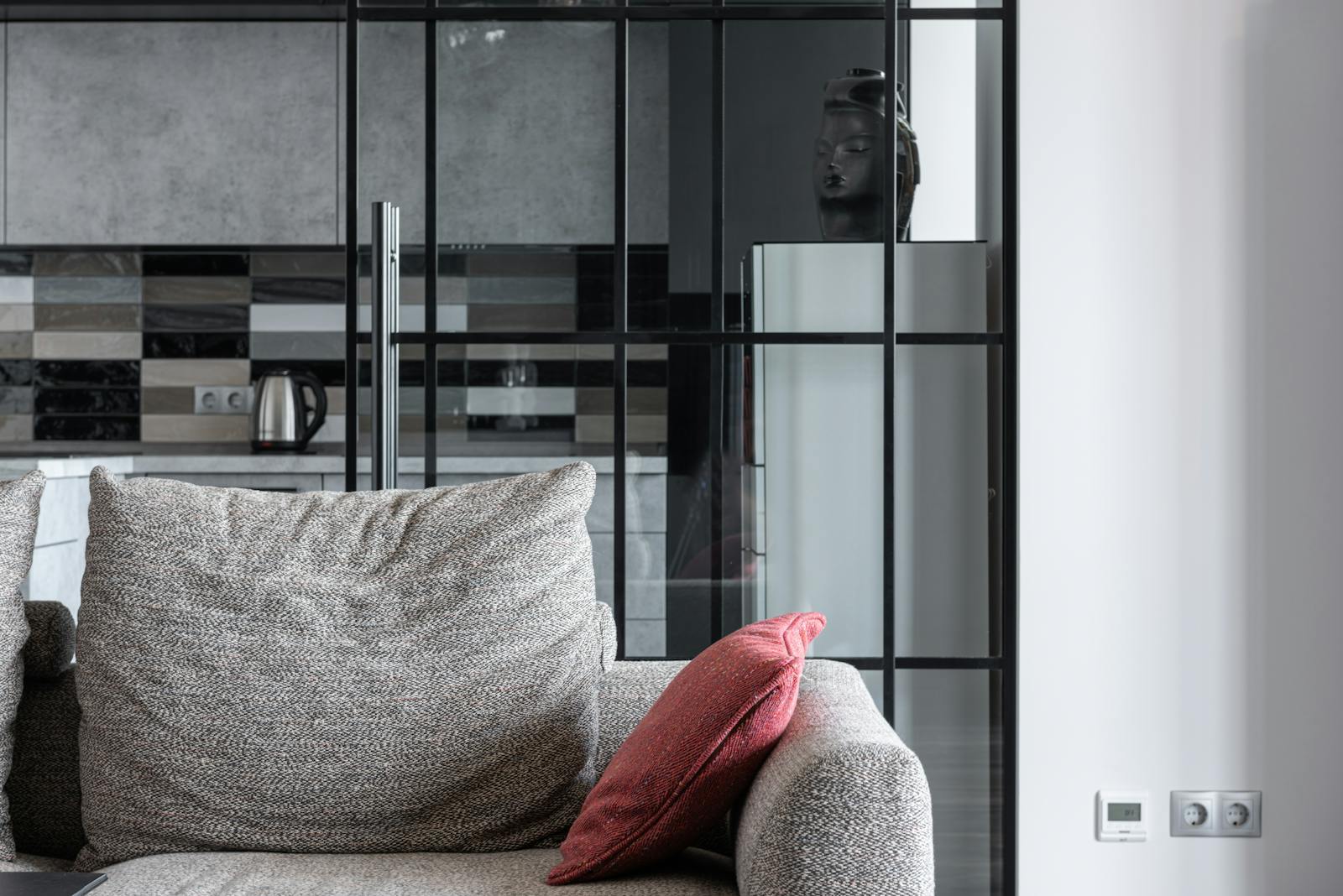 Salon confortable avec un canapé texturé avec un coussin rouge et une cuisine moderne en arrière-plan.